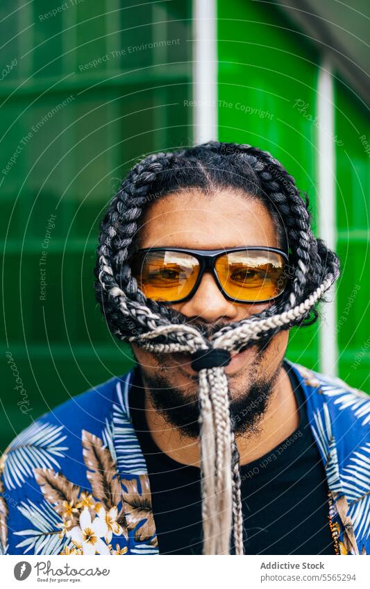 Smiling latin man with a knot of his hair braided in his face. braids sunglasses rope blue green background portrait beard shirt floral fashion close-up style