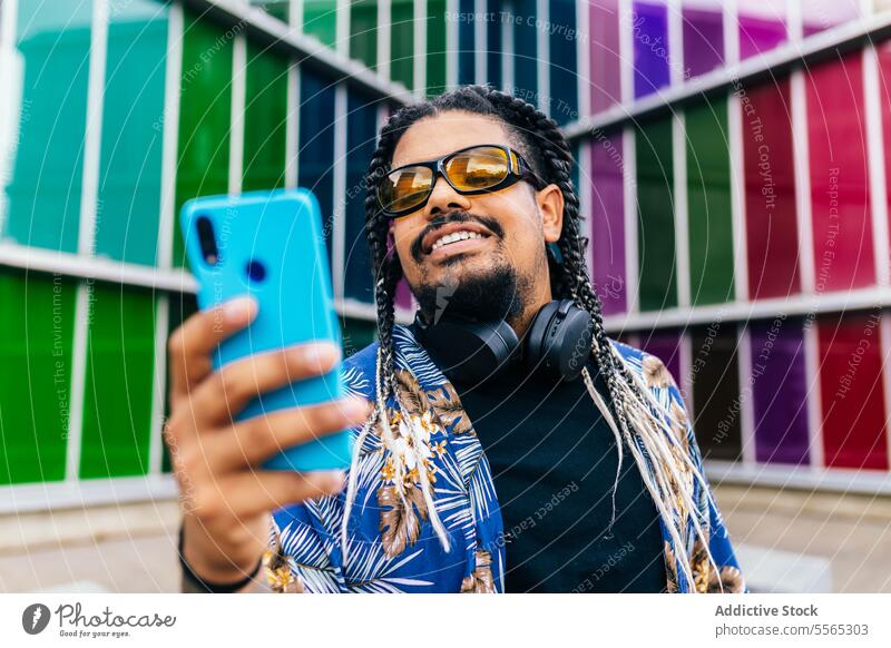 Latin man with headphones in his neck looking at blue smartphone in colorful urban place. braids smiling talking background sunglasses shirt tattoo outdoor
