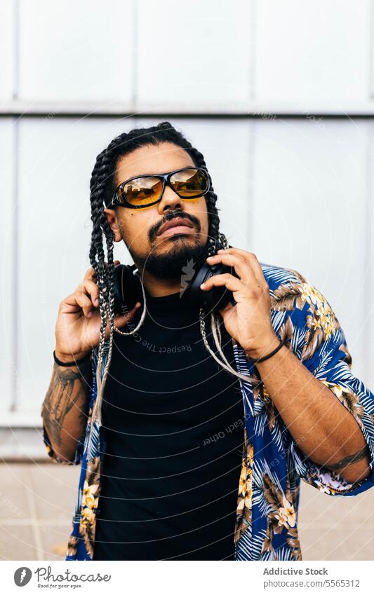 Latin man with braids and sunglasses looking away while holding his headphones around his neck. latin thought tattoo diversity listening vertical beard mustache