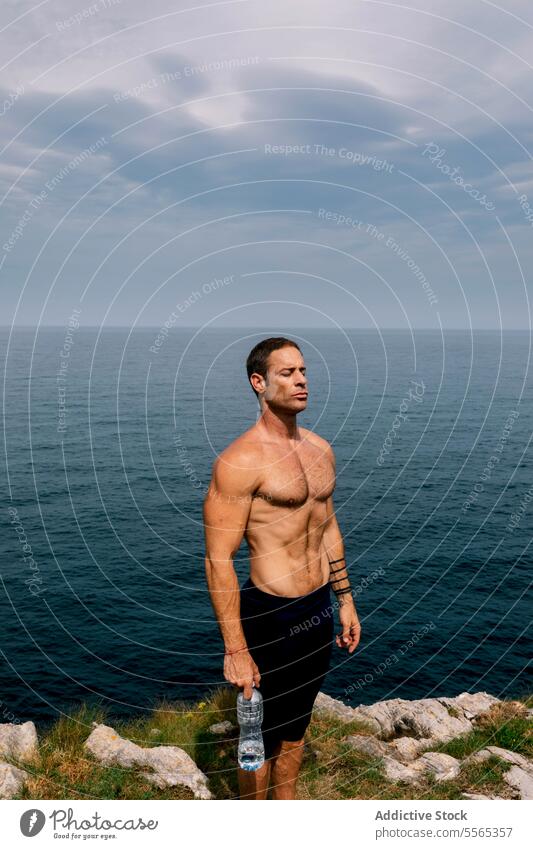 Man standing by the sea with water bottle Sea man toned physique sky clouds horizon fitness health nature outdoors workout hydration muscles serene calm coast