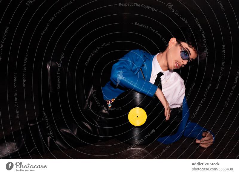 Retro-styled man lounging with vinyl record in the dark stylish glasses blue jacket leather pants retro music fashion 80s ambiance vintage youth subculture