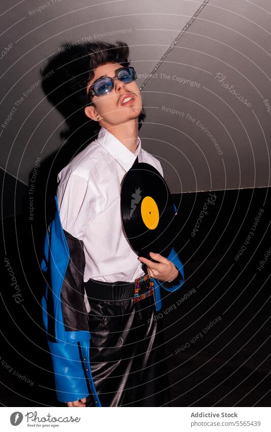 Retro-styled man with vinyl record in the dark stylish glasses lounging blue jacket leather pants retro music fashion 80s ambiance vintage youth subculture