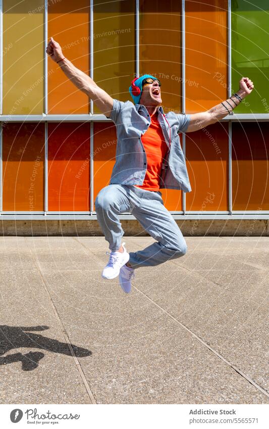Energetic man celebrates with leap by colorful backdrop jump celebration headphones music joy casual energy vibrant building architecture sunglasses excitement