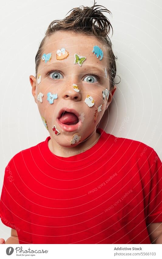 Boy with colorful stickers on his face and wet hair tongue animal playful child young expression red shirt white background fun silly emotion cheerful cute kid
