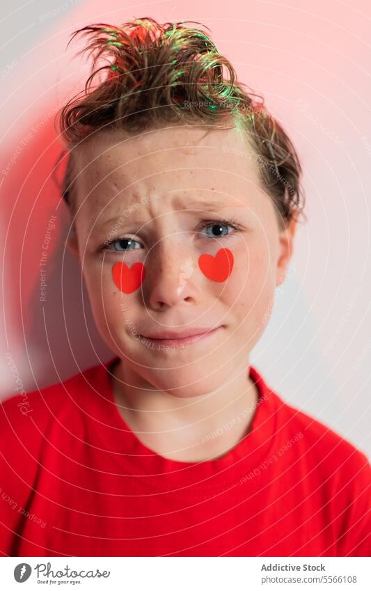 Calm boy with heart stickers on cheeks and abstract red patterns Boy curly hair calm gaze red shirt white background emotion child young expression love serene