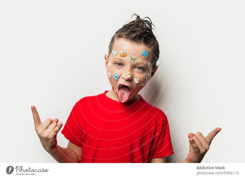 Boy with colorful stickers on his face and wet hair tongue animal playful child young expression red shirt white background fun silly emotion cheerful cute kid