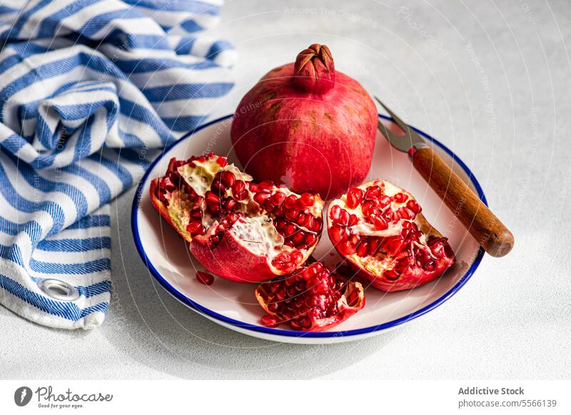 Open ripe pomegranate on plate with a rustic knife and a striped napkin. Pomegranate seed fruit red vibrant fresh cloth texture white blue background food juicy