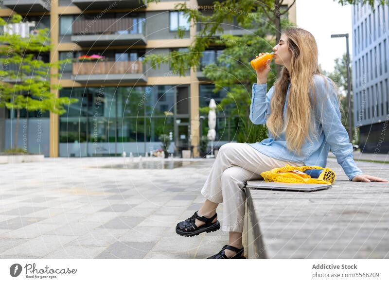 Female student sipping drink outdoors in lunchtime in city. blonde woman young gen-z drinking braces sitting juice caucasian blue shirt orange bottle urban