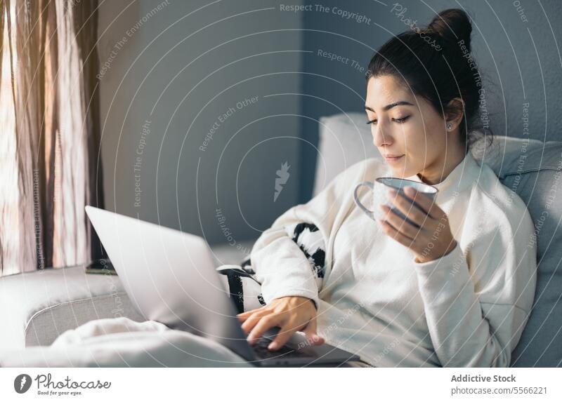 Crop young woman sitting with laptop and coffee cup using internet connection online living room at home female dark hair sofa gadget device beverage netbook