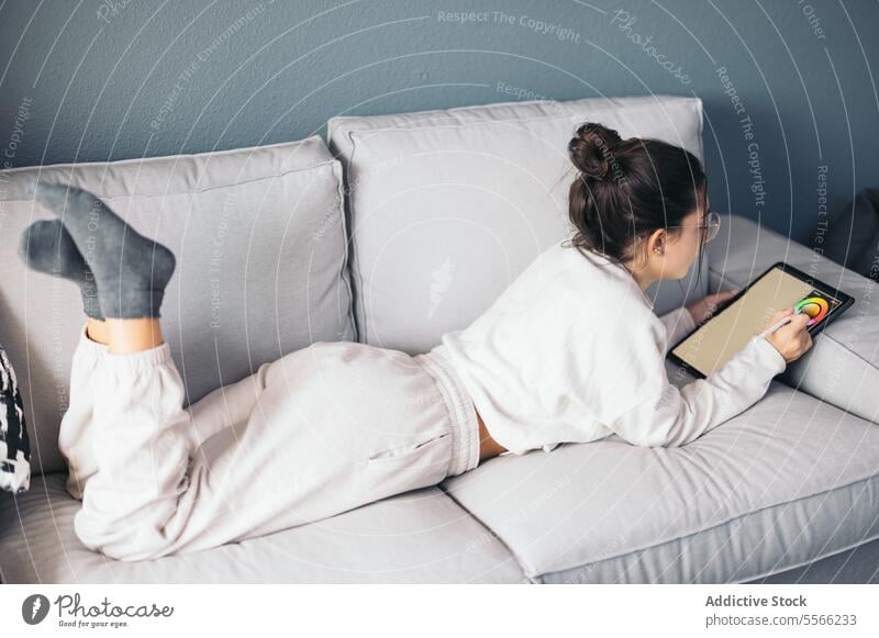 Young woman in eyeglasses lying on sofa and browsing digital tablet at home read e book cozy comfort online living room young female bookworm relax belly sock
