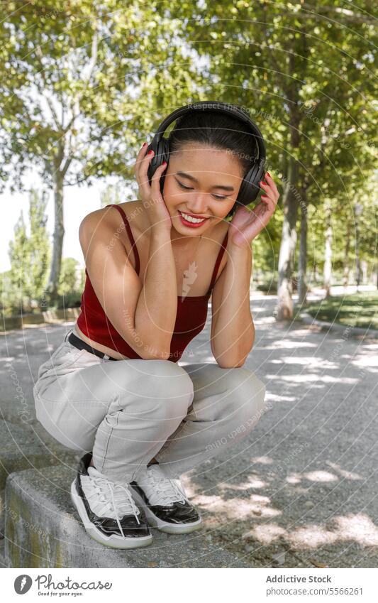 Asian Woman enjoying music in park woman headphones listening trees nature smile sunny day outfit summer relaxation outdoors leisure happiness style technology