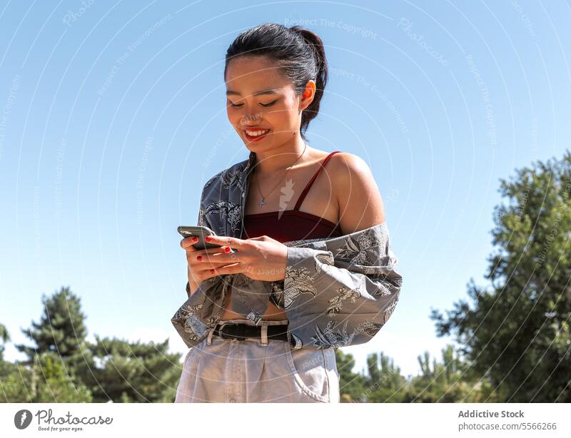 Asian woman Outdoor with smartphone use on a sunny day. outdoors stylish outfit mobile technology young female park summer modern casual messaging texting