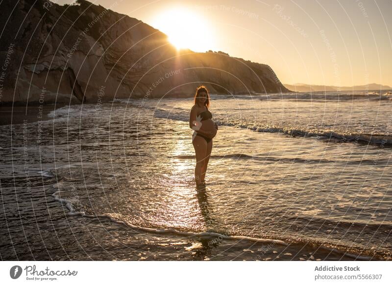 Smiling pregnant woman standing in sea water belly expect bikini summer sunset await touch female happy cheerful smile prenatal anticipate pregnancy maternal
