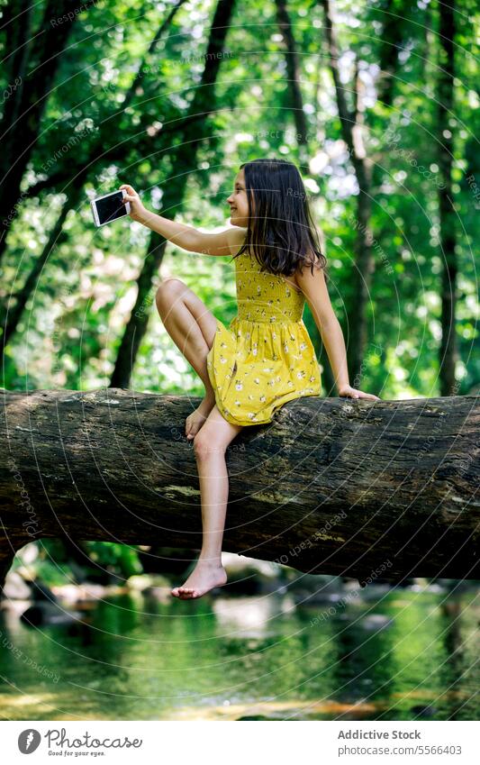 Preteen girl sitting on tree trunk while taking selfie forest break recreation nature adventure tranquil enjoy summer harmony holiday trip tourism young