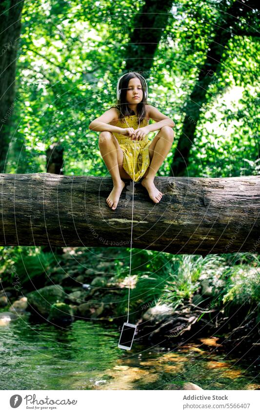 Preteen girl sitting on tree trunk while listening music on headphones with hanging mobile phone forest break recreation nature adventure tranquil enjoy summer