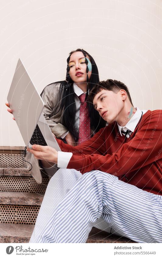 Two young fashion models sitting on urban stairs and looking at laptop. Work concept. gen-z woman work makeup tie shirt red eyes steps professional youth seated