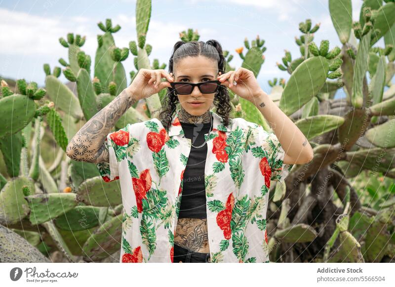 Tattooed woman standing on blurred cactus while adjusting sunglasses stylish nature tattoo attractive young fashion female pretty style cool beauty trendy