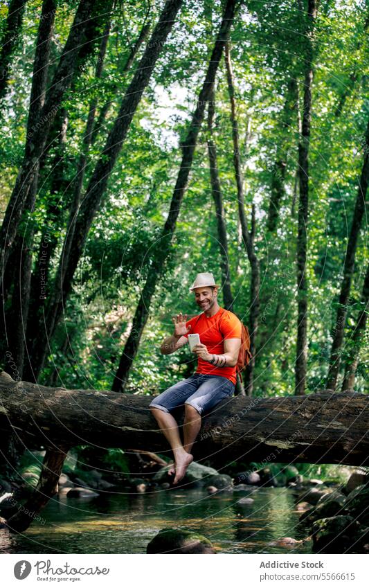 Man with hat and red t-shirt in conversation with smartphone amidst forest tranquility hiker man log talk tree green seated call technology communication