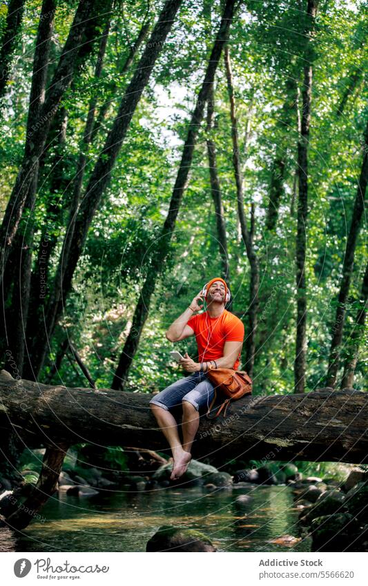 Man with cap sitting on a fallen tree trunk listening music over a serene forest stream. hiker headphones smartphone creek lay stretch man orange shirt nature
