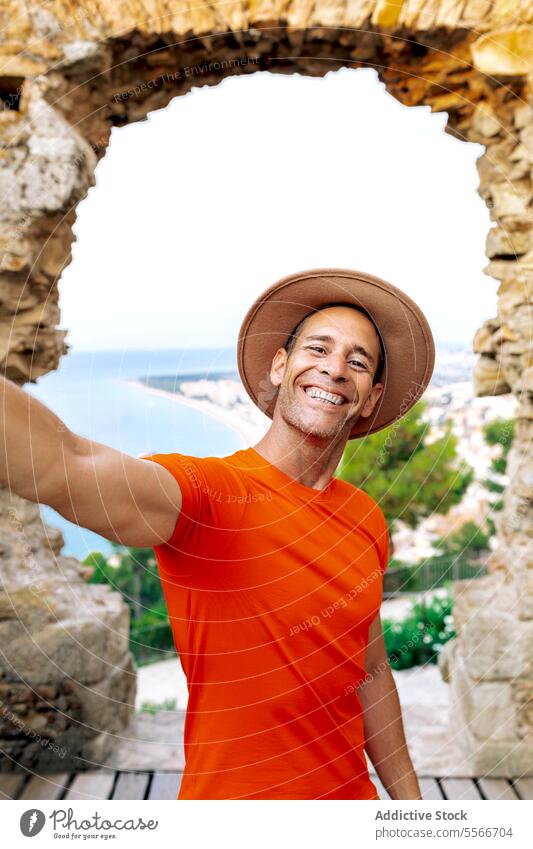 Happy traveler captures selfie against seaside Barcelona background. man phone camera smartphone joy looking arch coast orange shirt smile hat view stone