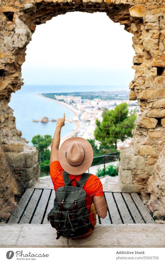 Traveler gestures towards seaside town from historic arch in Barcelona city. traveler stone man coast point tourist orange vertical holidays shirt hat backpack