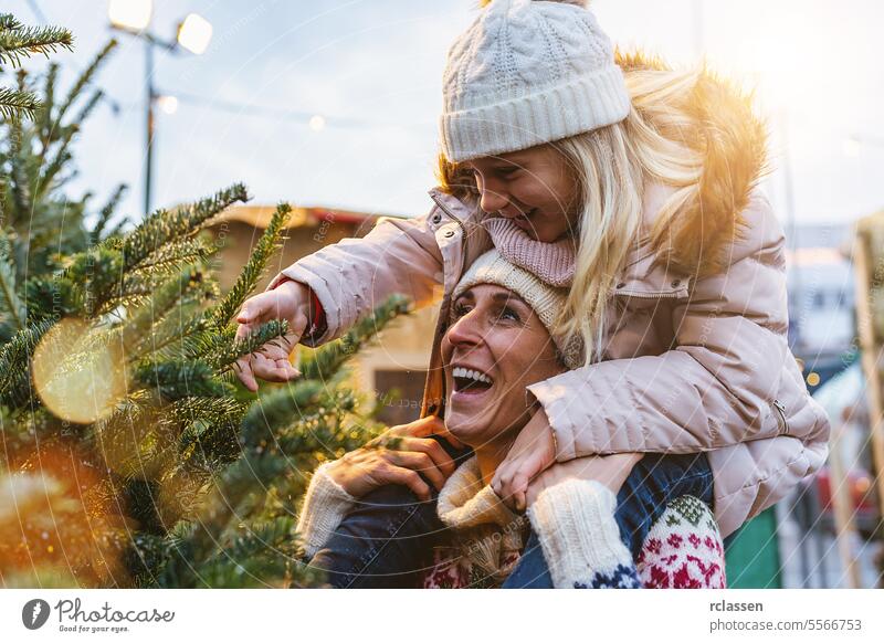 happy mother carries her little daughter piggyback and choosing Christmas tree at street market child surprised family skeptical redhead tradition choice smile
