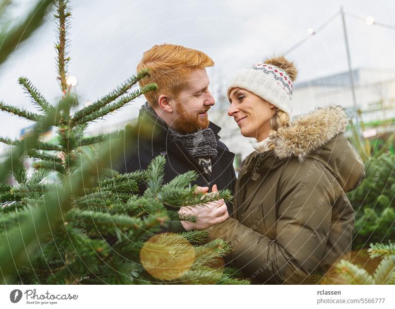 Couple in love at a Christmas tree market look at kiss family happy redhead tradition choice smile man love couple purchase farm pine tree norway shop buying