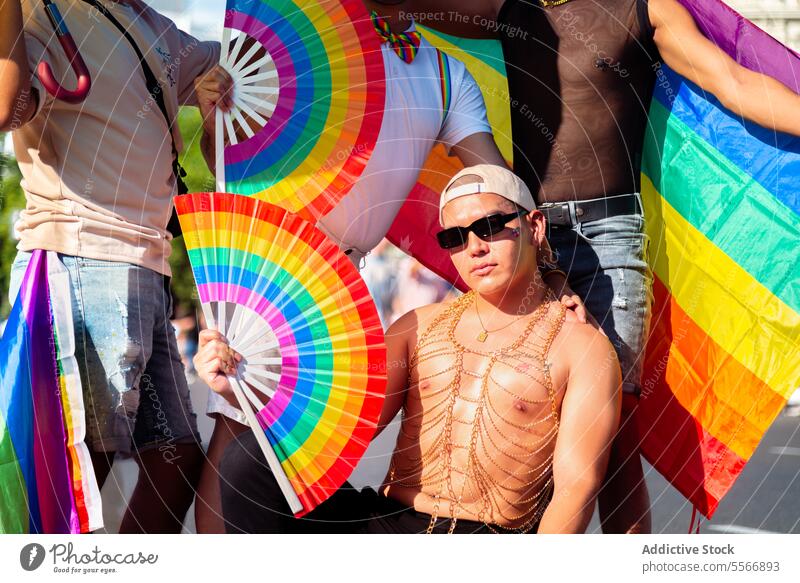 Confident man with paper fan at LGBT pride event Man sunglasses gold necklace pose flag sunlight vibrant color accessory confidence celebration festival