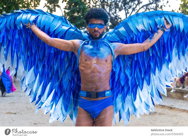 Black blue winged standing outdoors during LGBT pride event man tree confident sunglasses mystical paint vibrant adorn costume festival celebration feathers