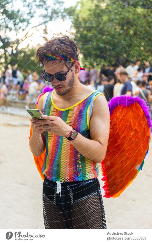 Man using phone at pride celebration in a park young accessories headband tank mobile festival sunglasses feather wing people event festive colorful wristband