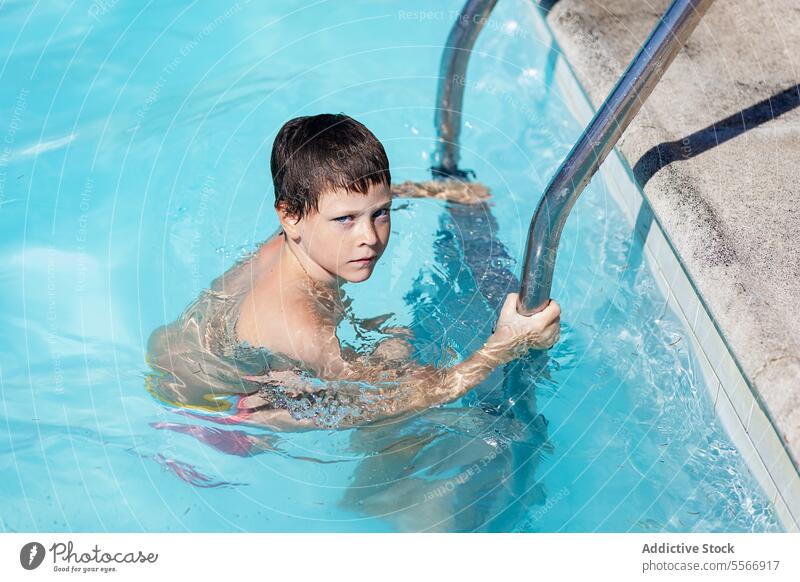 Boy in a pool gripping ladder with intent gaze. water summer swim blue child portrait hold metal clear outdoor fun leisure step handrail safety swimmer youth