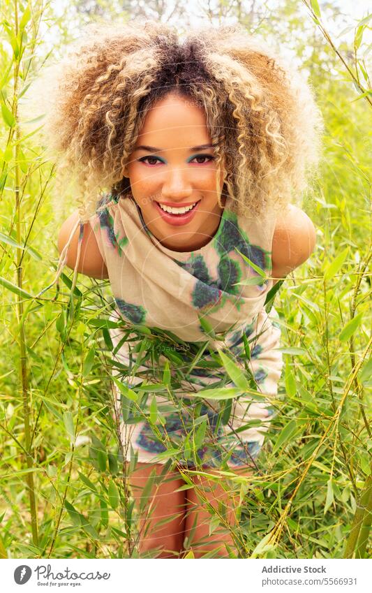 Ethnic woman amidst green reeds in patterned dress Woman blonde curly hair playful mysterious sleeveless nature camera gaze field outdoors environment tall look