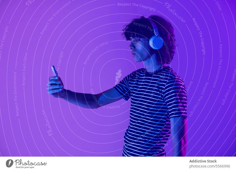 Latin man in headphones illuminated in purple viewing smartphone curly hair striped shirt latin gaze light screen focus technology fashion model electronic