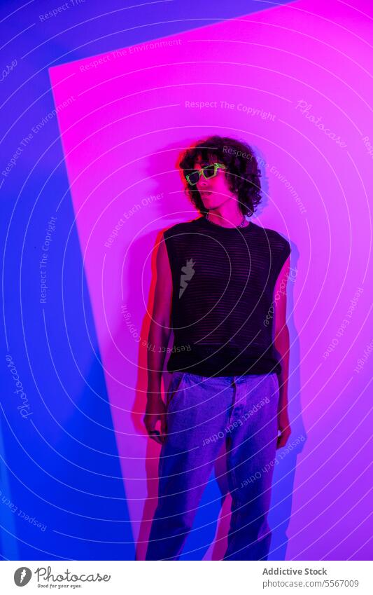 Curly-haired latin man in mesh tank with neon background curly style blue pink sunglasses top fashion pose light studio trendy modern cool vibe contrast color