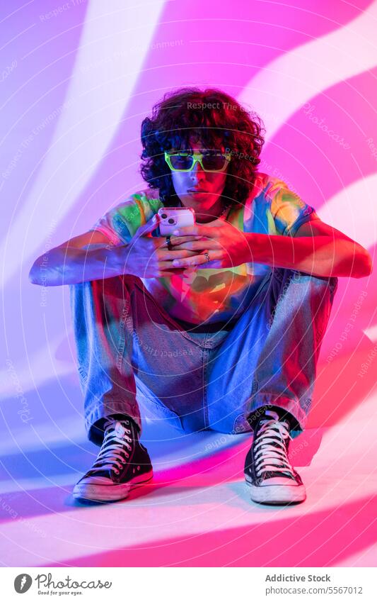 Engrossed latin man in tie-dye on phone with pink hue curly hair sit shirt smartphone use gradient backdrop fashion style jeans sneakers striped model pattern