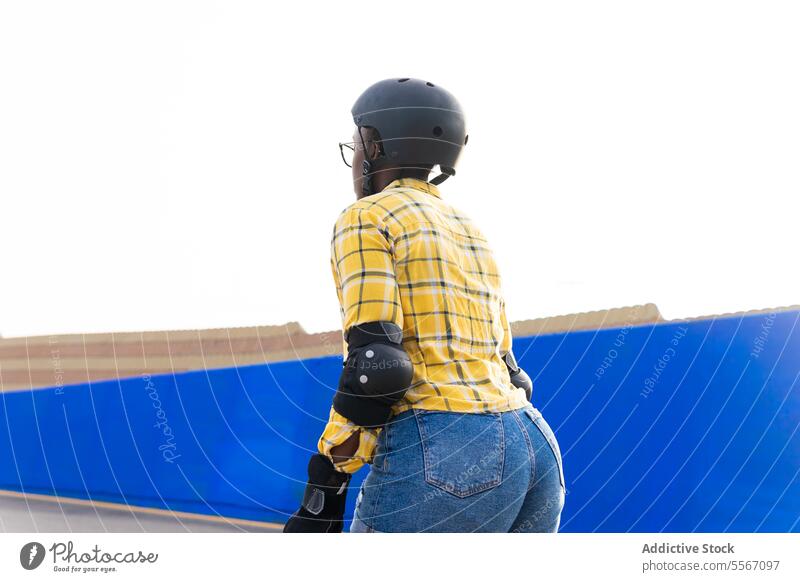 Woman In Protective Gear Roller Skating At Skate Park Against Clear sky Helmet Shirt Protection Blue Wall Sunny Casual Plaid Sport Leisure Weekend Practice Rink