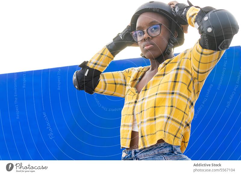 Thoughtful Woman Adjusting Helmet Against Blue Wall Eyeglasses Looking Away Sunlight Protection Gear Standing Casual Yellow Plaid Shirt Skate Park Clear Sky
