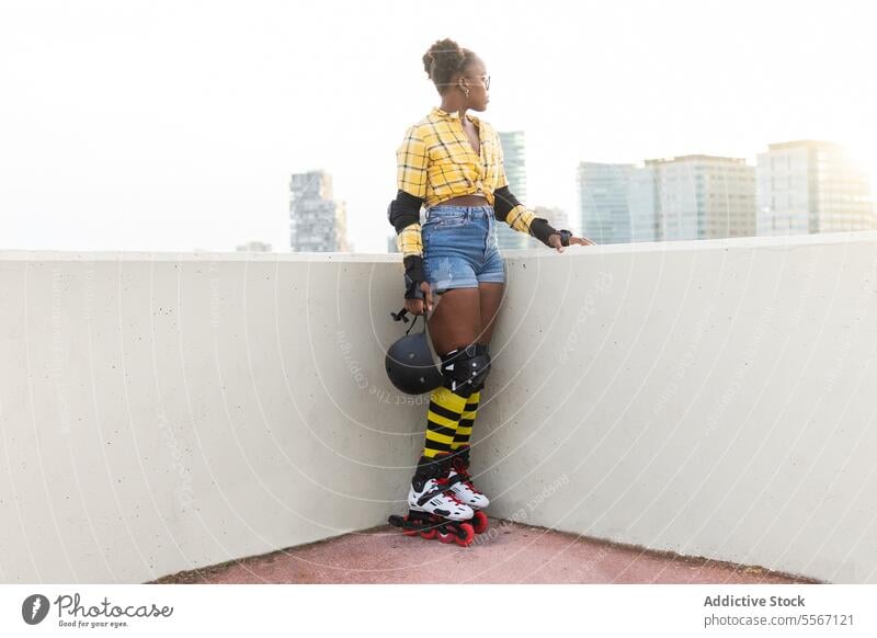 Thoughtful Woman In Roller Skates Holding Helmet At Skate Park Looking Away Wall Sunlight Protection Gear Standing Casual Shirt Shorts Sport Leisure Weekend