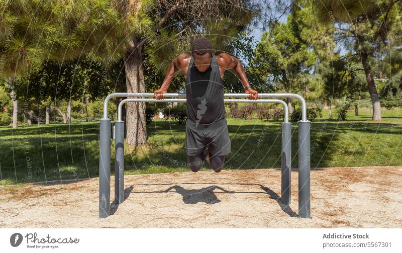 Black man in athletic wear performs a parallel bar dip exercise in a park with lush trees in the background outdoor strength training fitness workout grip sport