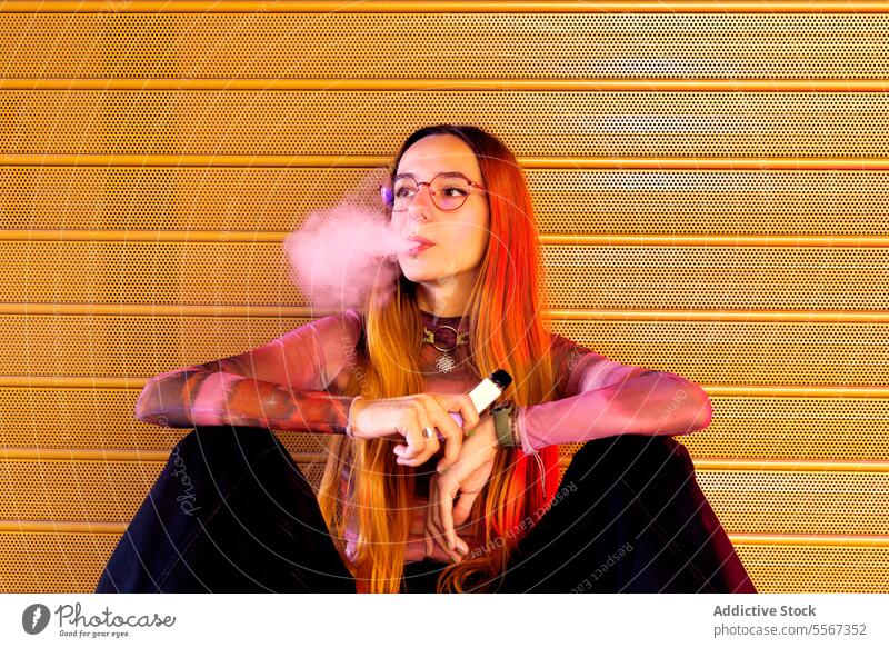 Young woman vapor exhales against orange backdrop gradient hair glasses purple background perforated e-cigarette contemplative pose style fashion seated