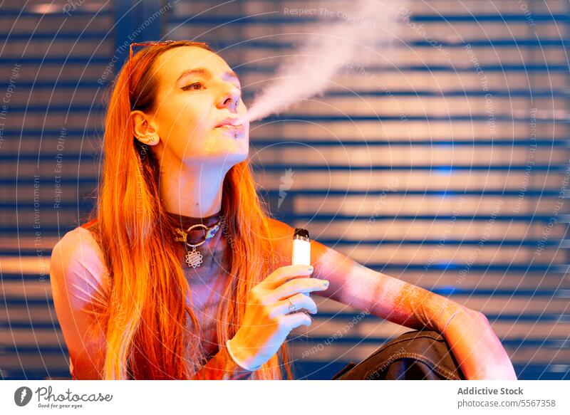 Young woman vapor exhales near blue metallic backdrop hair vaping e-cigarette background orange illumination ambient light city urban evening portrait