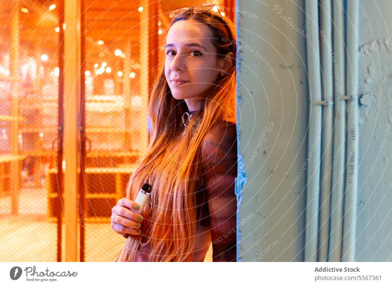 Young woman holding an e-cigarette against serene urban backdrop hair orange mesh fence teal wall metal pipe standing gaze poise young city texture gradient