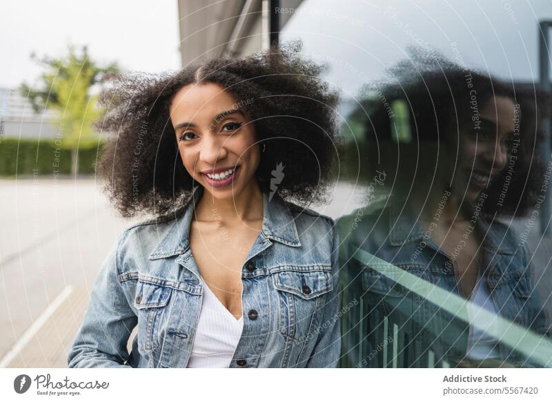Ethnic young woman smiling and leaning against a reflective glass pane curly hair denim jacket white top smile reflection joy street urban fashion casual style