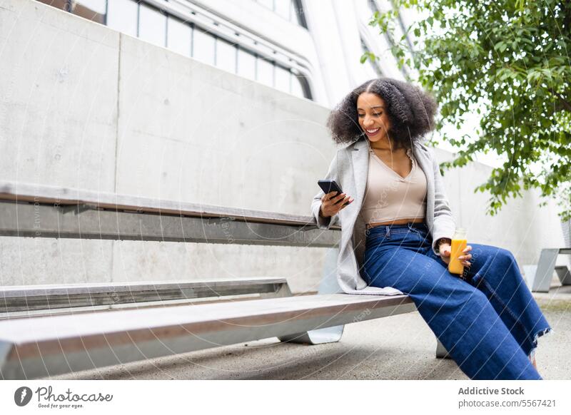 Ethnic woman with smartphone and juice in street curly hair outdoor modern building hand bottle drink reading browsing message sit urban refreshment break