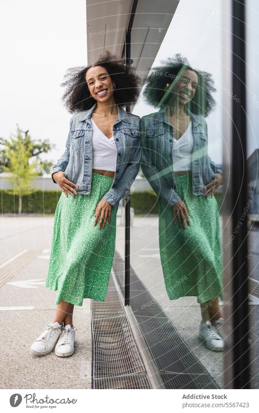 Ethnic young woman smiling and leaning against a reflective glass pane curly hair denim jacket white top green skirt smile reflection joy street urban fashion