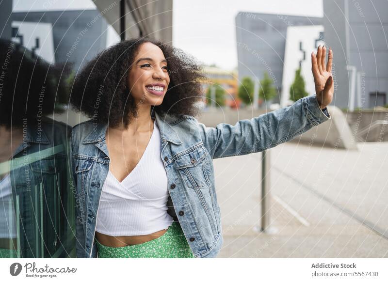 Ethnic young woman smiling and waving curly hair wave hello denim jacket white top green skirt reflection glass building cheerful modern urban smile greeting
