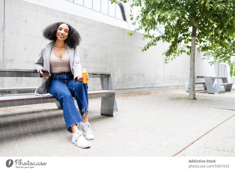 Ethnic woman with smartphone and juice in street curly hair outdoor modern building hand bottle drink browsing sit urban refreshment break leisure city