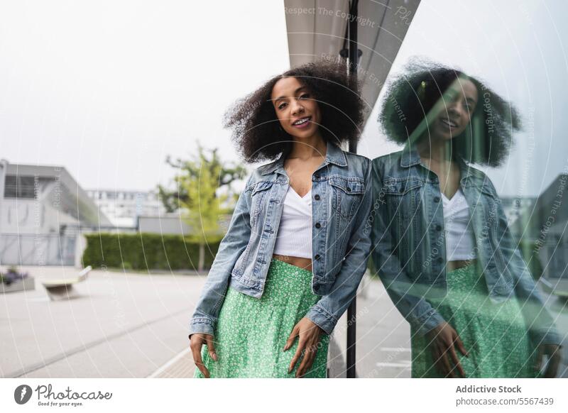 Ethnic young woman smiling and leaning against a reflective glass pane curly hair denim jacket white top green skirt smile reflection joy street urban fashion