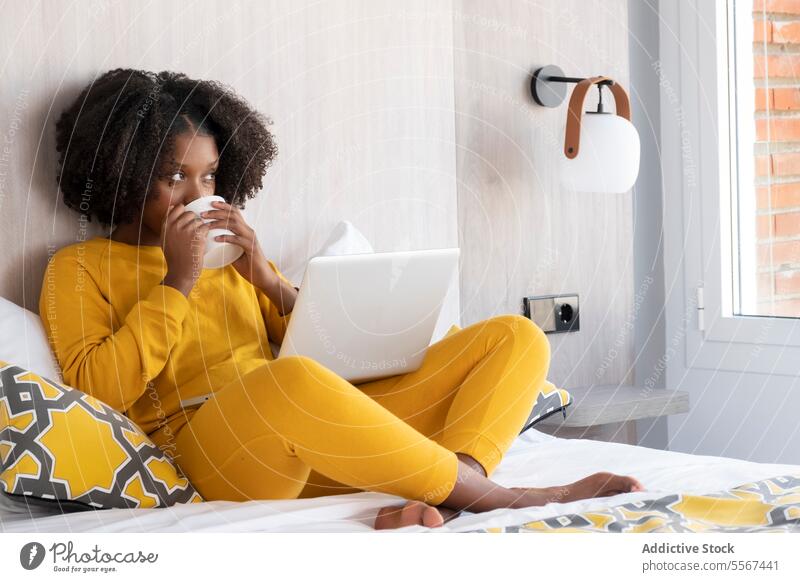 Black woman in cozy work moment at home yellow coffee laptop bed interior modern focus cushion wall lamp window brick relax technology device task online browse