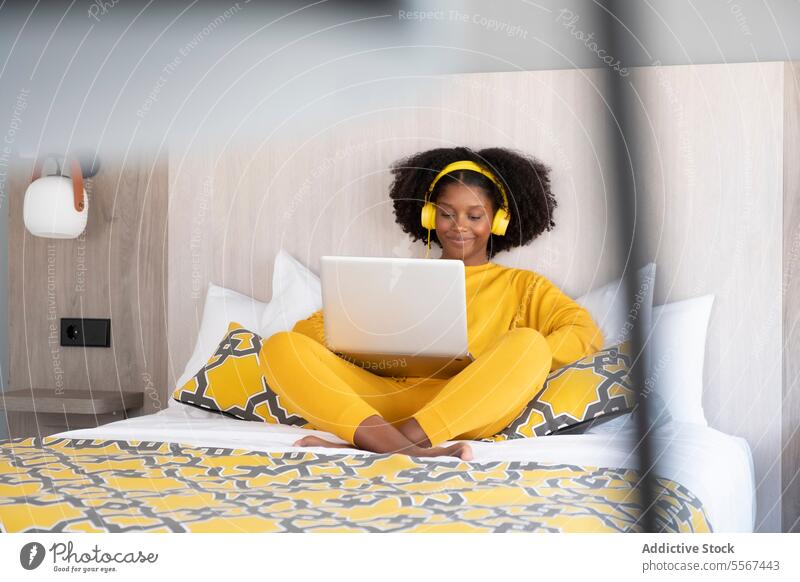 Black woman in digital relaxation in modern bedroom yellow laptop headphone enjoy female design outfit smile cushion pattern wall lamp sit technology music